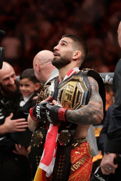 Ilia Topuria wearing two UFC championship belts over his shoulder, looking up during UFC 317.