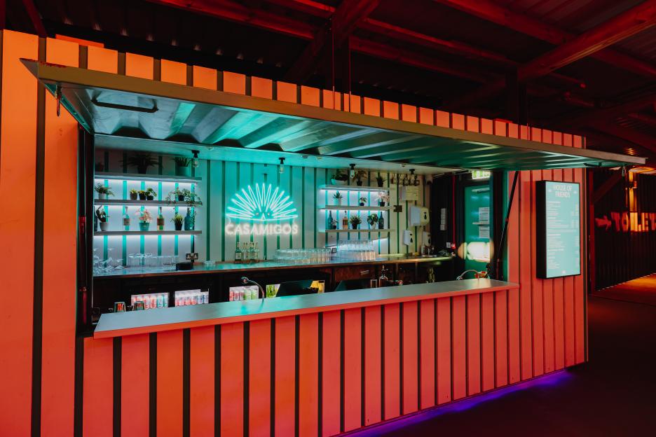 A rooftop bar illuminated with blue and red lights, featuring a "CASAMIGOS" neon sign and a menu on the right.