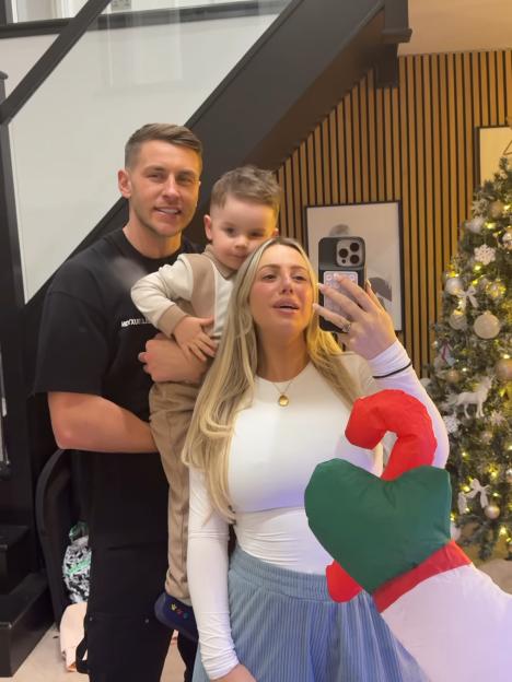Holly Hagan, her husband, and son posing for a selfie in front of a Christmas tree, holding an inflatable candy cane.