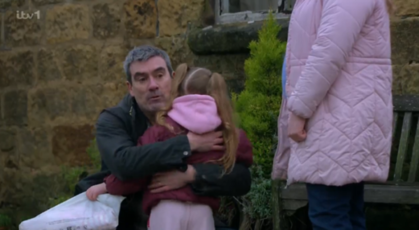 A man in a black jacket hugging a young girl in a pink hooded jacket, with a woman in a pink puffer coat standing to their right.